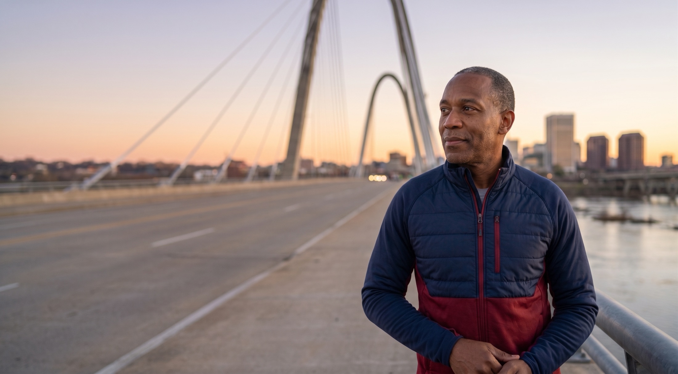 A man in his sixties pausing on a Richmond bridge at sunset, the city skyline behind him